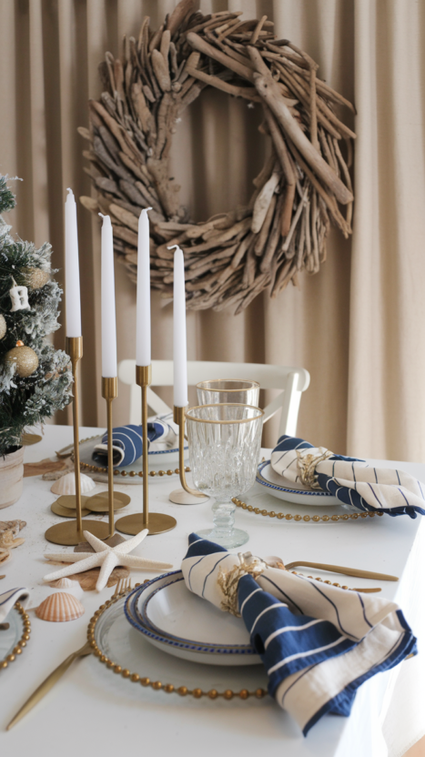 Coastal chic Christmas table setting with nautical elements, gold accents, and a driftwood wreath for a stylish holiday gathering.